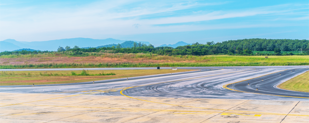 An empty airport runway and taxiway curving into the distance on a clear day, with grassy fields and trees in the background.
