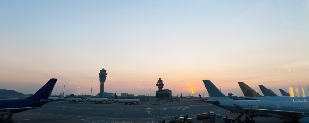 A wide view of an airport tarmac at sunset, showing airplane tails, the control tower, and airfield lighting glowing in the dusk.